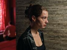 A woman in front of a wall, looking determined while a man is half-naked on a bed in the background (still 1)