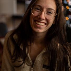 Delaney (woman with long brown hair and clear framed glasses) smiles to the camera in a headshot style, close up photo.