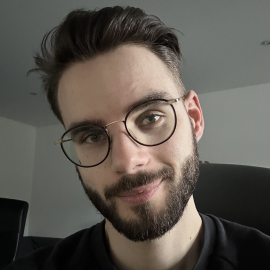 Photo de profil d'un homme blanc d'une trentaine d'années, avec des cheveux bruns, portant la barbe et des lunettes, souriant.